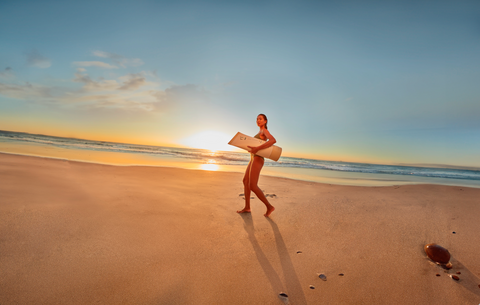 Person holding the Liquid Gold SPF50 Broad Spectrum Sunscreen tube as a surfboard on a beach at sunset